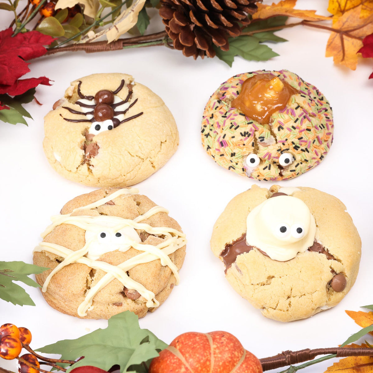 Four decorated chocolate chip cookies arranged on a white surface with autumn-themed props including pumpkins, pinecones, and fall leaves. The cookies are styled as a spider, a donut with caramel apple and sprinkles, a mummy with white drizzle, and a ghost with a marshmallow.